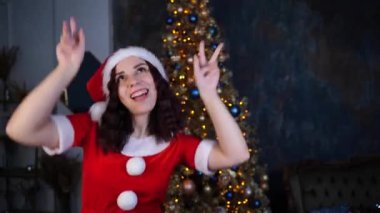 Noel Baba kostümlü genç bir kadın Noel ağacının arkasında dans ediyor. Mutlu bayan dinleniyor ve Noel 'i evde kutluyor..