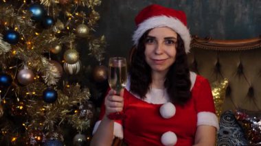 Noel Baba kostümlü genç bir kadın, elinde bir bardak şampanya, Noel ağacının yanındaki sandalyede oturuyor. Çekici kadın gülümsüyor, kameraya bakıyor.. 