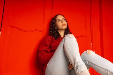 Genç bir kadın yerde oturuyor. Kıvırcık esmer, kırmızı duvarın yanında duruyor.