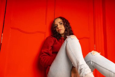 Genç bir kadın yerde oturuyor. Kıvırcık esmer, kırmızı duvarın yanında duruyor.