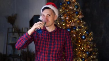 Noel Baba şapkalı genç adam karanlık odada Noel ağacının yanında şampanya içiyor. Erkek gülümsüyor, kameraya bakıyor. Evde Noel kutlaması kavramı