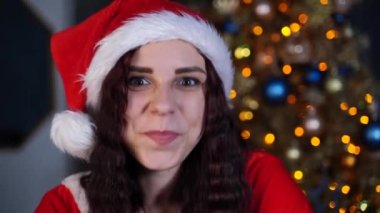 Noel Baba şapkalı çekici esmerin portresi Noel ağacının arka planında. Genç ve çekici bir kadın kameraya bakıp gülümsüyor. Evde Noel kutlaması kavramı.