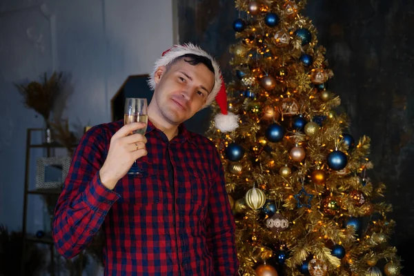 クリスマスツリーを背景にシャンパンのガラスとサンタの帽子の若い男 装飾的な装飾と針葉樹の木にポーズをとる大人の男性 家庭でのクリスマスのお祝いの概念 — ストック写真