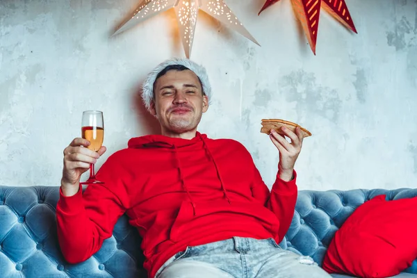 戴着桑塔帽的年轻人喝着香槟 吃着披萨 坐在房间的沙发上 快乐的男性休息与食物和酒精 在家里庆祝圣诞节的概念 — 图库照片