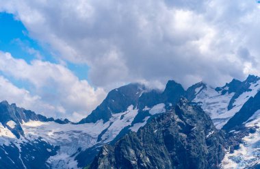 Bulutlu gökyüzüne karşı karlı dağ. Beyaz bulutların altından mavi gökyüzünde yüzen dağların üzerinden doğada güneşli bir günde karla kaplı..