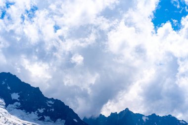 Bulutlu gökyüzüne karşı karlı dağ. Beyaz bulutların altından mavi gökyüzünde yüzen dağların üzerinden doğada güneşli bir günde karla kaplı..