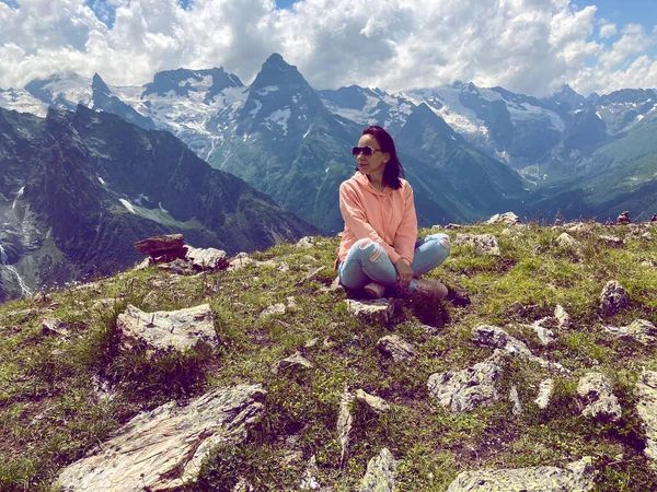 Young woman in sunglasses sitting on mountain top in sunny weather. Female tourist posing for camera on background of amazing mountain landscape in summertime