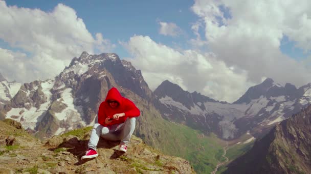 Junger Mann sitzt auf einem Felsen und macht mit dem Smartphone Fotos von der Berglandschaft. Männliche Reisende nutzen Handy gegen bewölkten Himmel an sonnigem Tag in den Bergen. — Stockvideo