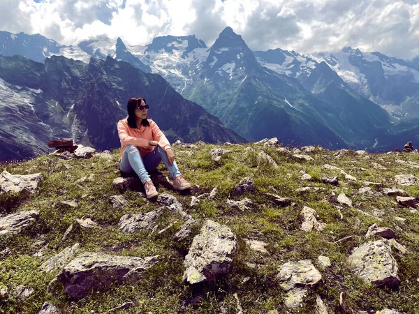 Young woman in sunglasses sitting on mountain top in sunny weather. Female tourist posing for camera on background of amazing mountain landscape in summertime