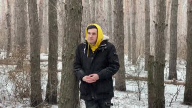 Sarı başlıklı ve siyah ceketli genç adam kış ormanlarında yürürken esniyor. Yetişkin erkek etrafına bakıyor, karlı havada ağaçların arasında duruyor..