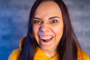 Suratı asık genç bir kadının portresi. Sarı kapüşonlu çılgın kadın, gri tuğlalı arka planda maymunluk yapıyor.