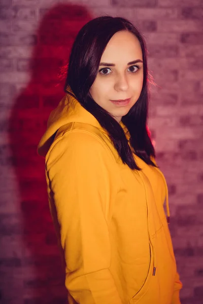 A young woman in a fashionable yellow hoodie. Portrait of a beautiful brunette in a yellow hoodie against a brick wall.