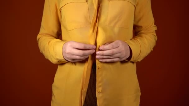 มุมมองด้านหน้าของร่างกายของหญิงสาวในเสื้อสีเหลืองที่ไม่มีปุ่ม ปิดขึ้นของผู้หญิงที่จําไม่ได้ ปุ่มเสื้อของเธอบนพื้นหลังสีส้ม . — วีดีโอสต็อก
