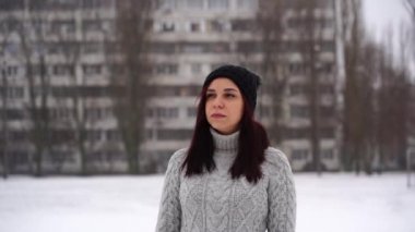 Gri örgü süveter ve şapkalı düşünceli genç bir kadın kış mevsiminde sokakta dikiliyor. Güzel esmer bakışlı ve bulutlu havada bir şeyi yansıtıyor..