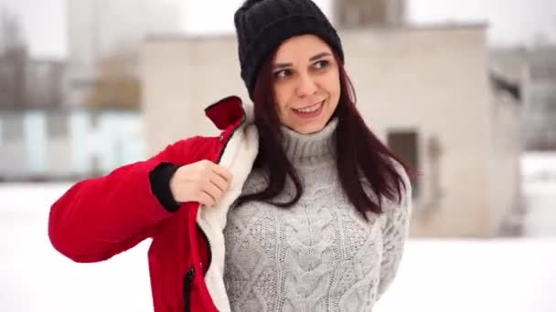 一个戴着帽子的年轻女子站在街上 穿着红色夹克 在寒冷的天气里 成年黑发人在冬季散步时暖和起来 — 图库视频影像