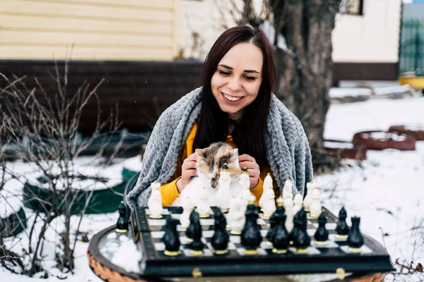 Young woman sitting with cat and playing chess in yard. Female wrapped in grey plaid sitting on street with pet and playing in board game in winter season