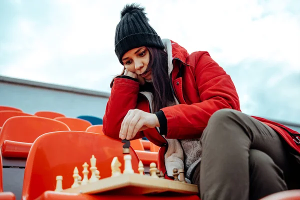 Young woman in winter clothes plays chess, sitting on stadium bleachers alone. Female in black cap with chess on sports stadium in cloudy weather