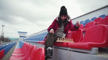 Kışlık elbiseli genç kadın satranç oynuyor, stadyum tribünlerinde tek başına oturuyor. Siyah şapkalı kadın, bulutlu havada spor stadyumunda satranç oynuyor.