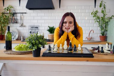 Mutfak masasında satranç oynayan genç bir kadın. Kadın mantıksal bir masa oyununda kendisiyle birlikte mutfakta oynuyor.