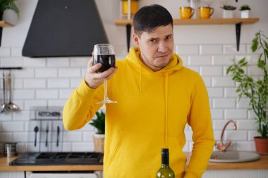 Rahatlamış genç adam, mutfak masasında duran kırmızı şarap kadehiyle poz veriyor. Yetişkin adam mutfakta alkolle dinleniyor..