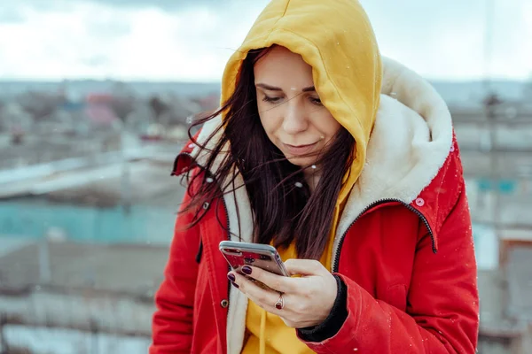 Portrait of young woman with mobile phone on roof of building. Brunette in yellow hoodie and red jacket browsing smartphone in cloudy weather