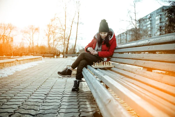 Young woman in winter clothes plays chess, sitting on bench in city park. Female in black cap with chess on wooden pew in cloudy weather