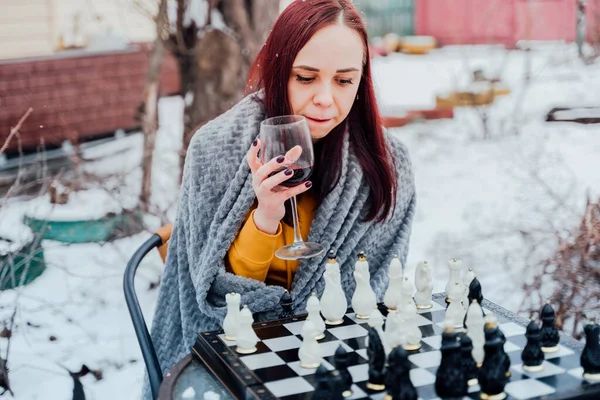 Young woman playing chess in yard. Female wrapped in grey plaid sitting on street playing in board game in winter season