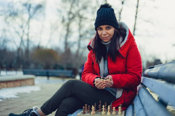 Young woman in winter clothes plays chess, sitting on bench in city park. Female in black cap with chess on wooden pew in cloudy weather