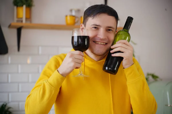 Relaxed young man poses with bottle and glass of red wine in kitchen. Adult happy guy presses alcohol to face