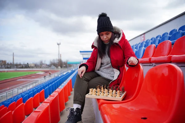 Young woman in winter clothes plays chess, sitting on stadium bleachers alone. Female in black cap with chess on sports stadium in cloudy weather