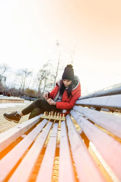 Young woman in winter clothes plays chess, sitting on bench in city park. Female in black cap with chess on wooden pew in cloudy weather