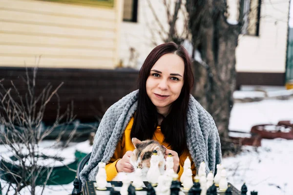 Young woman sitting with cat and playing chess in yard. Female wrapped in grey plaid sitting on street with pet and playing in board game in winter season