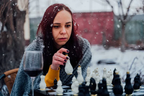 Young woman playing chess in yard. Female wrapped in grey plaid sitting on street playing in board game in winter season
