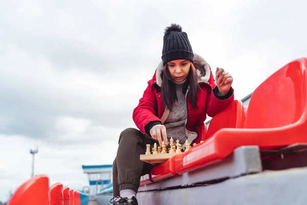 Young woman in winter clothes plays chess, sitting on stadium bleachers alone. Female in black cap with chess on sports stadium in cloudy weather