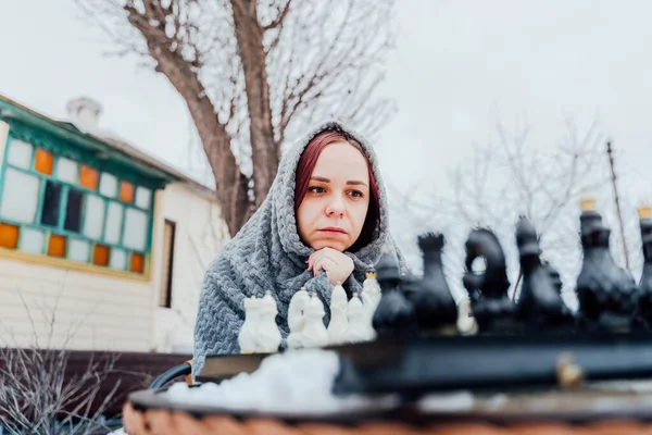 Young woman playing chess in yard. Female wrapped in grey plaid sitting on street playing in board game in winter season