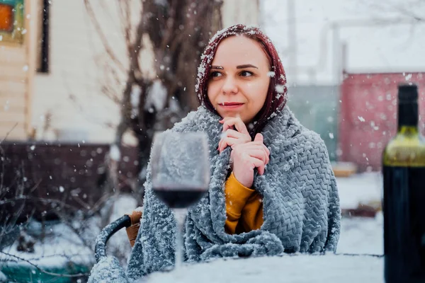 Young woman sitting in yard and drinking red wine in snowy weather. Female wrapped in grey plaid sitting on street with alcohol in winter season