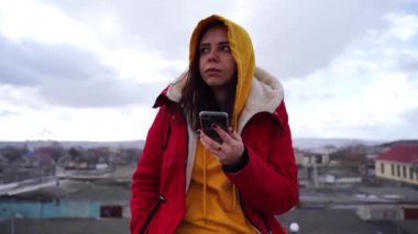 Binanın çatısında cep telefonu olan genç bir kadının portresi. Sarı kapüşonlu ve kırmızı ceketli esmer bulutlu havada akıllı telefona bakıyor..