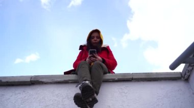 Cep telefonlu genç bir kadın beton korkulukta oturuyor. Sarı kapüşonlu ve kırmızı ceketli esmer bulutlu havada akıllı telefona bakıyor..