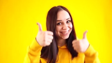 Sarı arka planda gülümseyen ve baş parmağını gösteren genç bir kadının portresi. Sarı kapüşonlu mutlu esmerin yaklaş. Onayını göster..