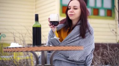 Bahçede oturan ve karlı havada kırmızı şarap içen genç bir kadın. Gri ekoseye sarılı kadın kış mevsiminde alkolle sokakta oturuyor..