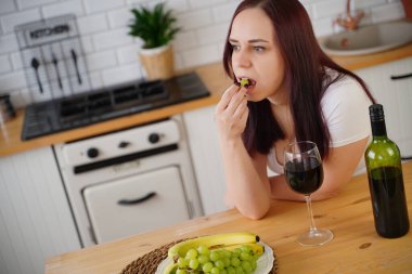 Genç bir kadın mutfakta yeşil üzüm yiyor. Yetişkin bir kadın hafta sonu meyve ve alkolle dinleniyor.