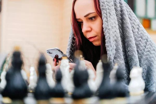 Young woman playing chess and browsing smartphone in yard. Female wrapped in grey plaid with mobile phone sitting on street playing in board game in winter season