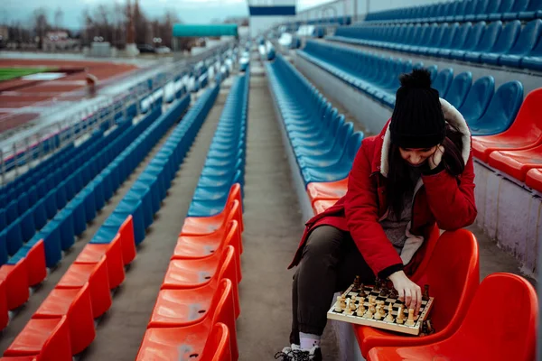 Young woman in winter clothes plays chess, sitting on stadium bleachers alone. Female in black cap with chess on sports stadium in cloudy weather