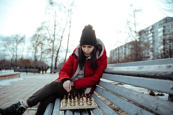 Young woman in winter clothes plays chess, sitting on bench in city park. Female in black cap with chess on wooden pew in cloudy weather