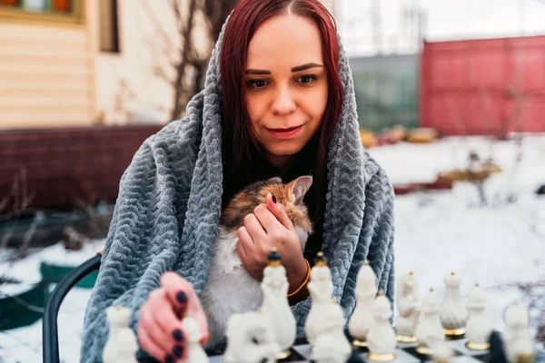 Young woman sitting with cat and playing chess in yard. Female wrapped in grey plaid sitting on street with pet and playing in board game in winter season