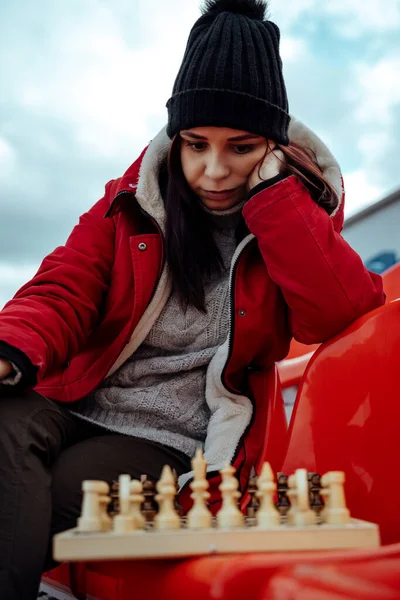 Young woman in winter clothes plays chess, sitting on stadium bleachers alone. Female in black cap with chess on sports stadium in cloudy weather