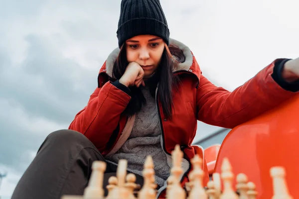 Young woman in winter clothes plays chess, sitting on stadium bleachers alone. Female in black cap with chess on sports stadium in cloudy weather