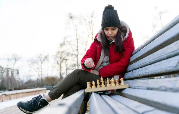 Young woman in winter clothes plays chess, sitting on bench in city park. Female in black cap with chess on wooden pew in cloudy weather