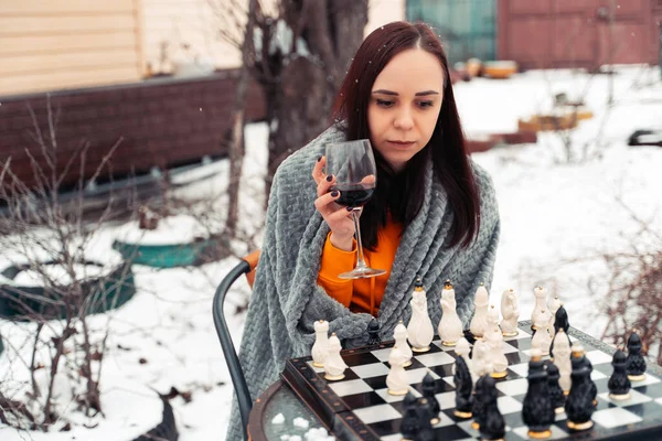 Young woman playing chess and drinking red wine in yard. Female wrapped in grey plaid with alcohol sitting on street playing in board game in winter season
