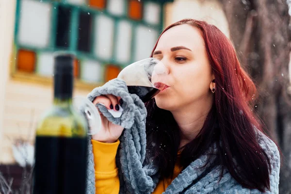 Young woman drinking red wine in yard. Female wrapped in grey plaid with alcohol sitting on street in winter season.